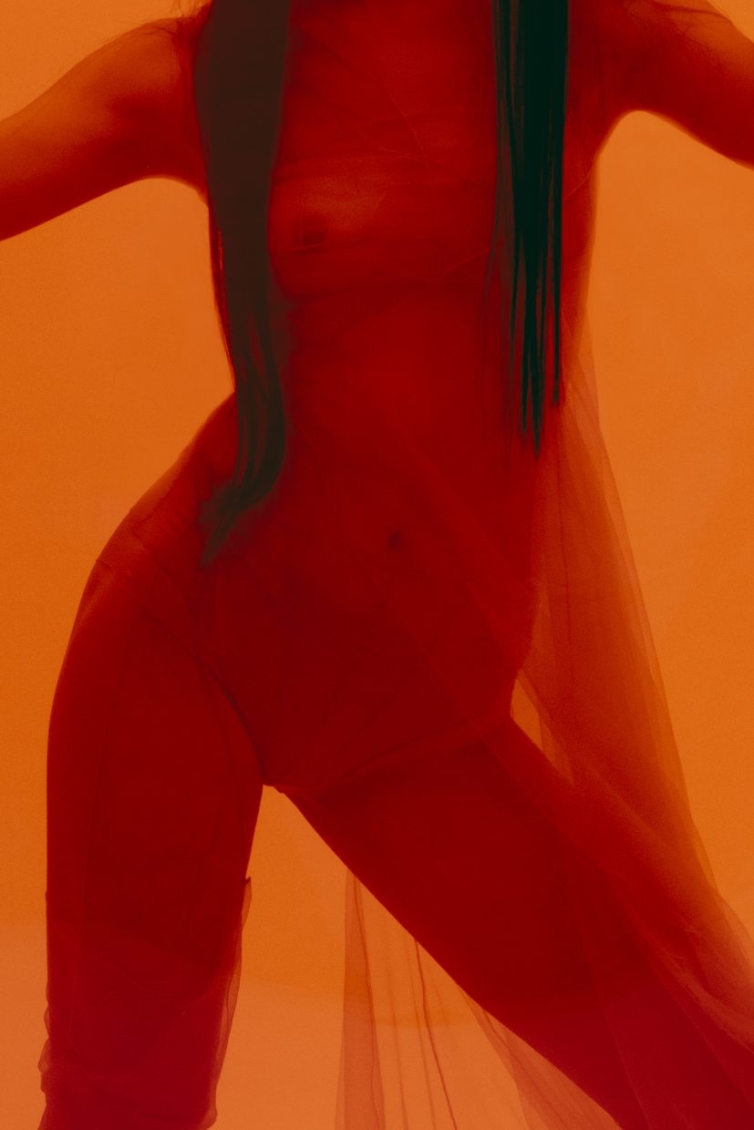 Das von Olga Urbanek vor einem orangefarbenen Hintergrund fotografierte Model Loo Loo trägt transparente Kleidung