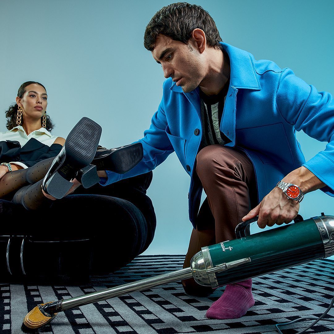 a man wearing a Rolex vacuums under the legs of a woman sitting on a sofa staring at him