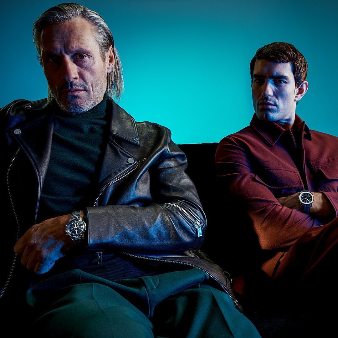 two men wearing Rolexes are sitting on a sofa on a shaded blue background