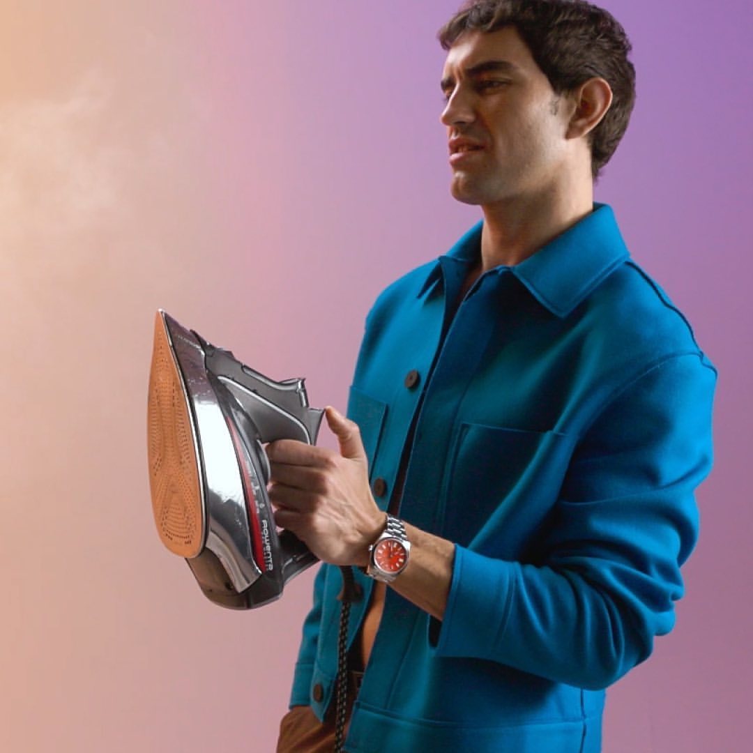 a man wearing a Rolex uses an iron on a pastel background ranging from pink to purple
